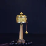 Tibetan prayer wheel with beaded tassel on wooden stand, showing engraved Om Mani Padme Hum mantras and brass hanging bell in front view