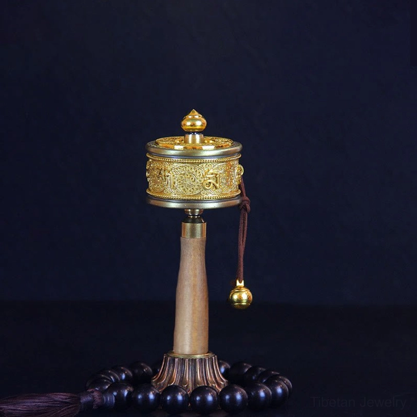 Tibetan prayer wheel with beaded tassel on wooden stand, showing engraved Om Mani Padme Hum mantras and brass hanging bell in front view