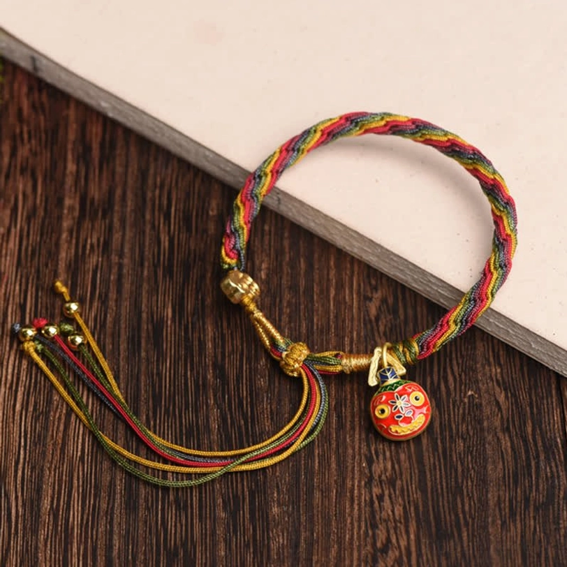 Tibetan prosperity bracelet with rainbow woven rope, gold fasteners, enamel charm, and beaded tassel on notebook