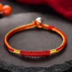 Handmade Tibetan red and gold braided protection bracelet with silver clasp, displayed on dark stone surface with spiritual ornament