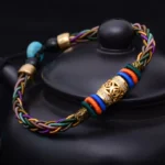 Tibetan eight knot copper coin red string bracelet handwoven close-up on black ceramic surface showing multicolor braided cord with gold and turquoise beads