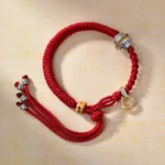 Tibetan red string braided bracelet with diamond knot, carved silver bead, and gold clasp on wooden background