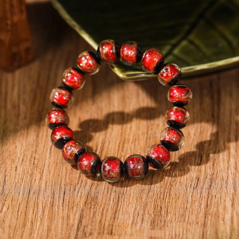 Single reddish-brown Tibetan liuli glass bead bracelet with uniform glossy beads and subtle metallic flecks, shown from overhead view on wooden surface with green leaf