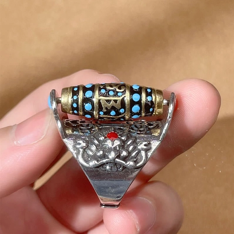 Tibetan rotating dzi ring held in finger showing retro silver base, central red inlay stone, smooth rotatable mantra bead design