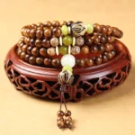 Tibetan rosewood mala bracelet with 108 beads displayed overhead on carved wooden stand, showing warm brown wood beads and decorative Bodhi seed accents with tassel