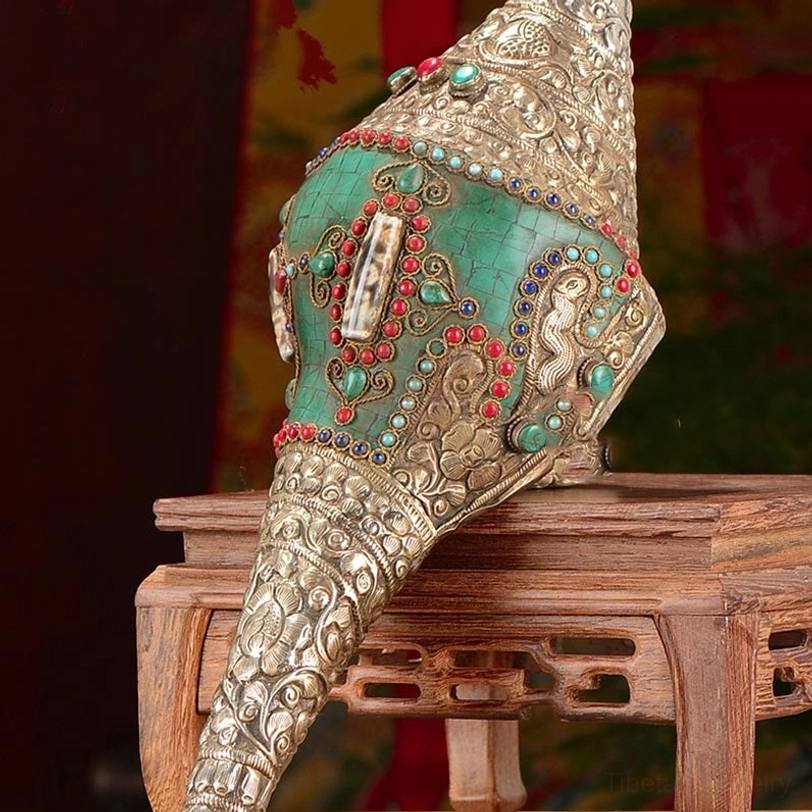 Tibetan decorated shankha conch shell close-up side view on wooden stand displaying intricate silver embossed motifs, turquoise and gemstone inlays