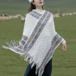 Off-white Tibetan rhombus knit tassel poncho shawl front three-quarter view on grassland with colorful ethnic border
