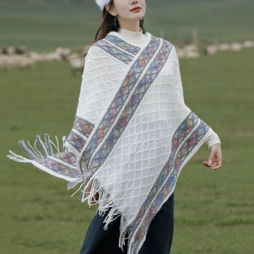 Off-white Tibetan rhombus knit tassel poncho shawl front three-quarter view on grassland with colorful ethnic border