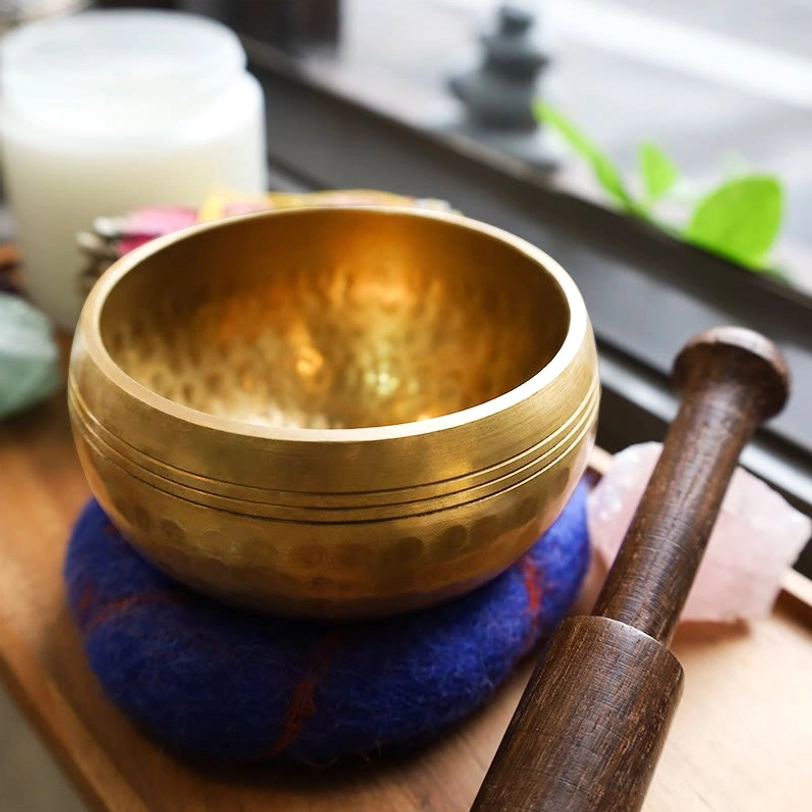 Tibetan brass singing bowl with wooden mallet and blue wool cushion on windowsill meditation setup