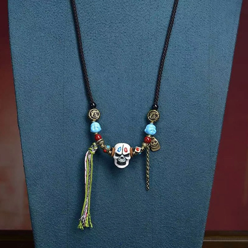Tibetan evil-protection skull necklace with black cord, blue gemstone beads, metal chain accent, and green woven tassel on dark blue background