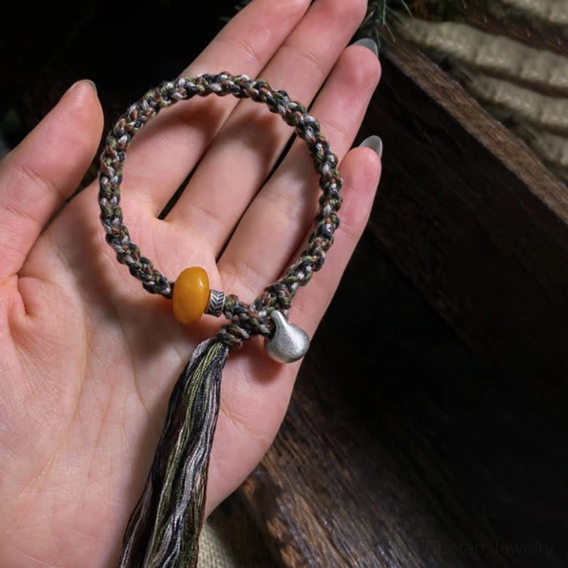 Handmade Tibetan protection bracelet held in palm on wooden desktop showing multicolor braided cord, golden amber bead, silver charm, and protective tassel