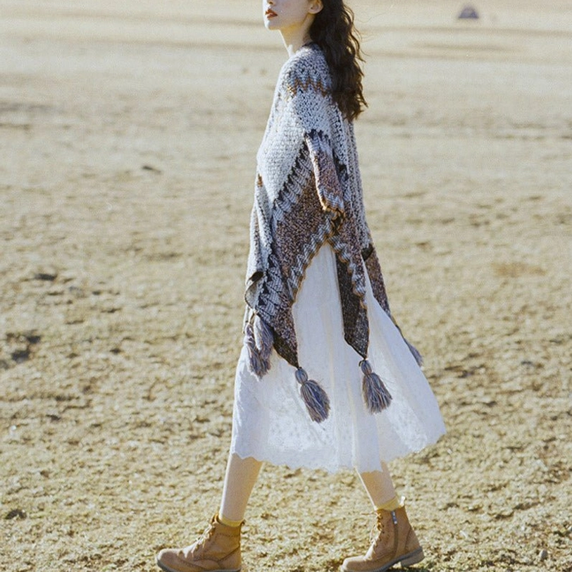 Side walking view of Tibetan striped poncho shawl with asymmetric hem and multiple tassel drops in neutral knit tones