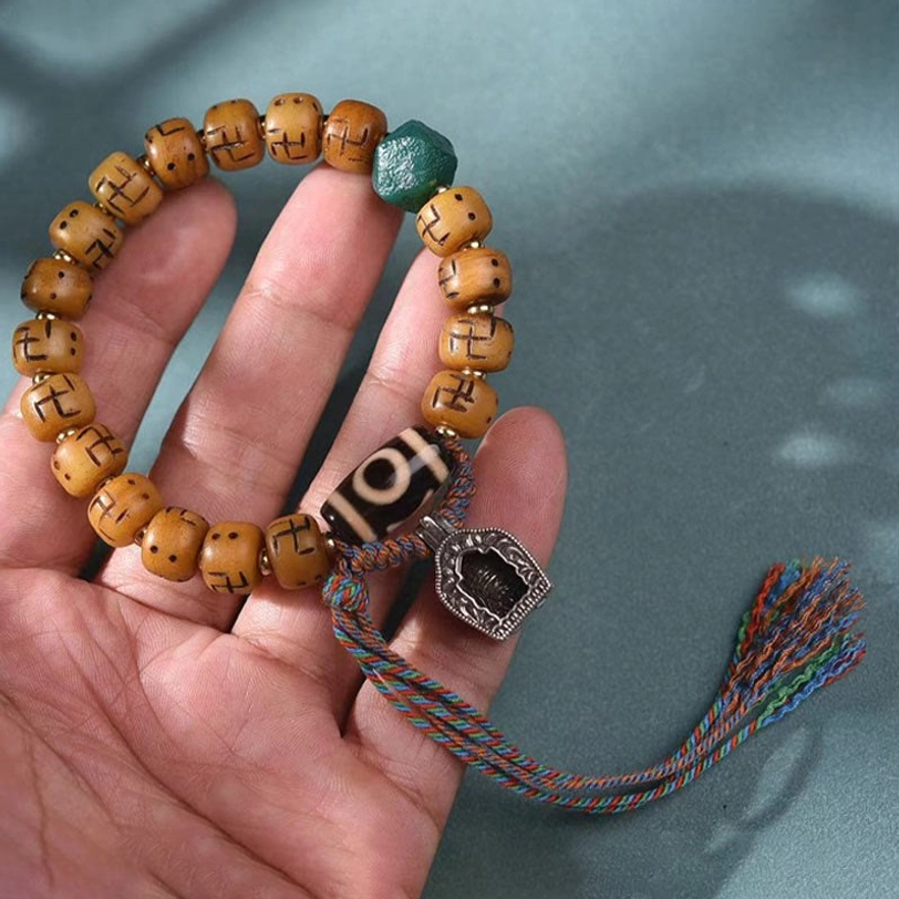 Close-up hand-held view of Tibetan camel bone bracelet showing carved swastika beads, black dzi bead, silver charm, and braided tassel detail