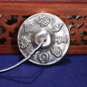 Single Tibetan tingsha bell eight auspicious symbols silver-toned copper leather strap front view blue fabric backdrop