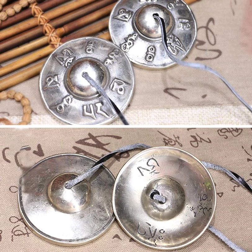 Pair of Tibetan tingsha bells white copper mixed embossed motifs leather straps oblique view bamboo mat cloth tabletop