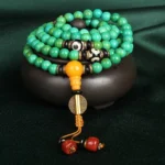 Tibetan turquoise 108 mala necklace with three-eye Dzi beads and amber guru bead, displayed on dark velvet with wooden base, top-down view