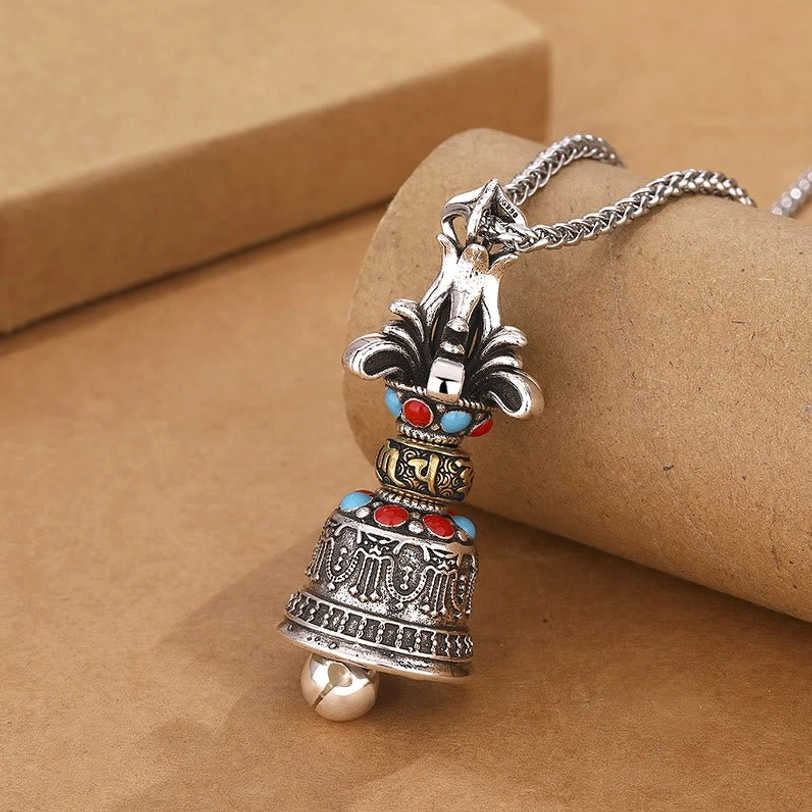 Tibetan Vajra Bell pendant necklace in vintage silver with red and blue gemstone inlays, featuring five-pronged dorje design with engraved Om Mani Padme Hum mantra, shown from front-left angle on kraft paper background