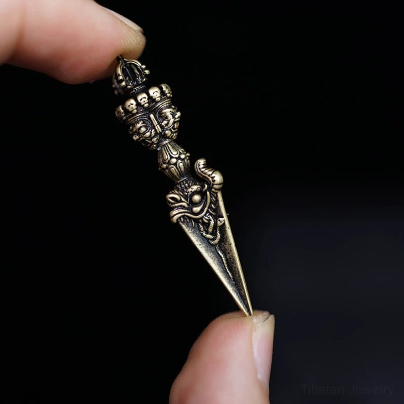 Handcrafted Tibetan Vajra phurba in dark antiqued bronze color held by thumb and index finger, religious carving patterns on dark background