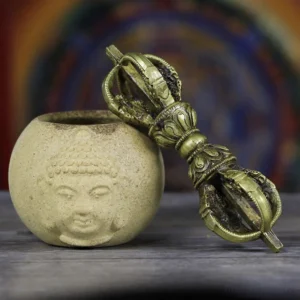 Handmade Tibetan five-prong vajra dorje in antique bronze copper finish, displayed on Buddha ceramic altar decoration, showing intricate carved metal texture and ritual details from oblique side angle