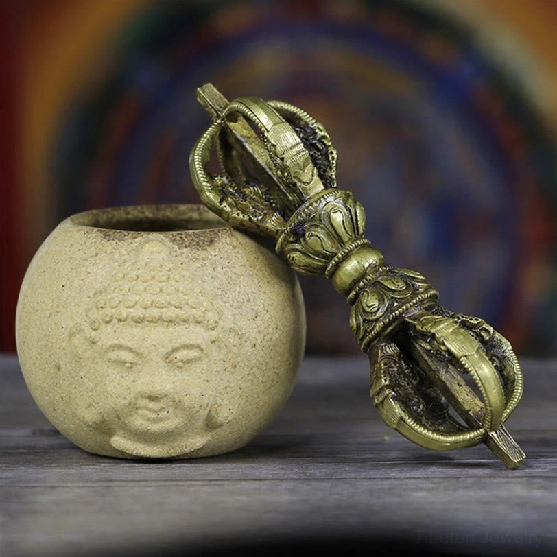 Handmade Tibetan five-prong vajra dorje in antique bronze copper finish, displayed on Buddha ceramic altar decoration, showing intricate carved metal texture and ritual details from oblique side angle