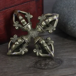 Tibetan double dorje vajra ritual object displayed from side angle, showing aged brass patina and ornate hollow prong carvings on wooden surface