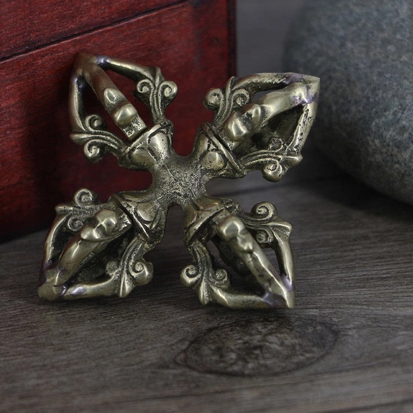 Tibetan double dorje vajra ritual object displayed from side angle, showing aged brass patina and ornate hollow prong carvings on wooden surface