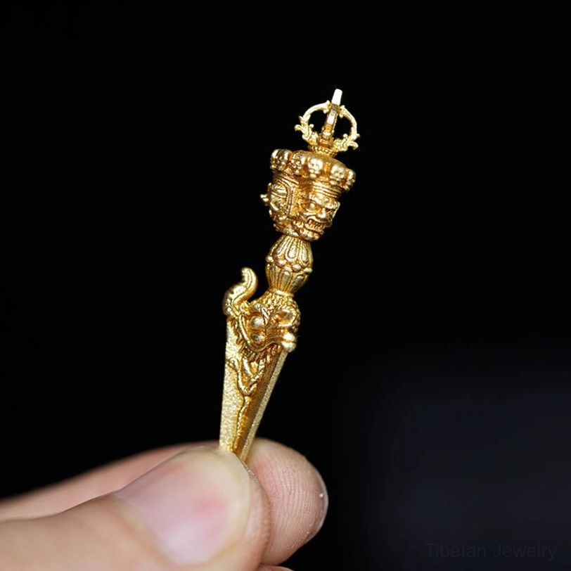 Handcrafted Tibetan Vajra pendant held by thumb, polished gold finish with detailed decorative engravings, pure dark background