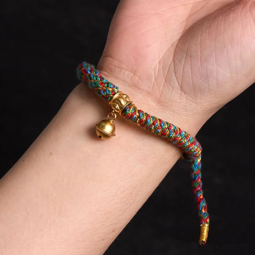 First-person wrist view of adjustable Tibetan woven bracelet with multicolor braid, gold bell charm, and long adjustable tail cord