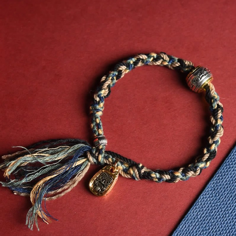 Handmade Tibetan Zakiram bracelet flat lay showing multicolor braided rope with carved gold charm and engraved silver barrel mantra bead