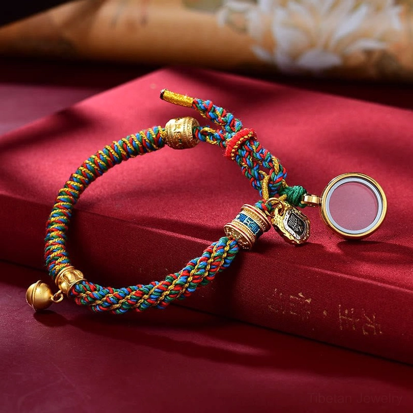 Tibetan multicolored woven bracelet with round empty locket pendant, gold bell charm, and Ghau prayer box compartment on burgundy velvet background