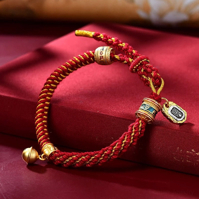 Tibetan red handwoven amulet bracelet with solid red cord, gold-toned Zakiram charm, bell pendant, and scripture tag on red velvet background