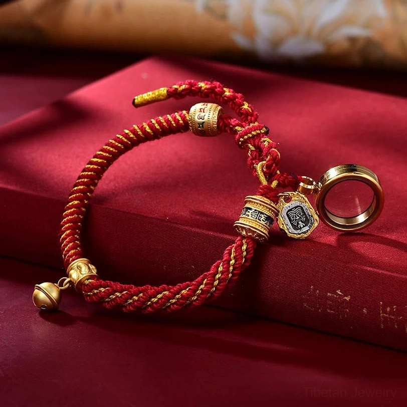 Tibetan red braided amulet bracelet with gold hardware, round Ghau prayer box locket pendant, scripture tag, and bell charm on burgundy background
