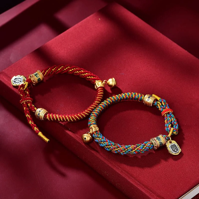 Two Tibetan amulet bracelets side by side—red and multicolored braided ropes with matching gold metal pendants and scripture charms on velvet