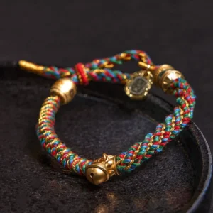 Close-up side view of handmade Tibetan Buddhist amulet bracelet featuring twisted rainbow cord, gold-tone hardware, and engraved charm beads