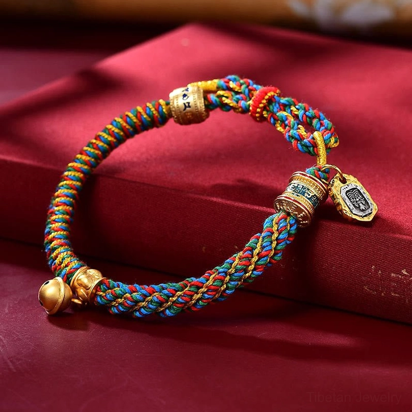Tibetan spiritual protection bracelet with blue-red braided rope and mixed metal charms on red silk fabric background