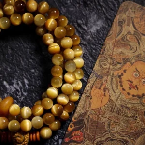 Close-up detail of golden Tiger Eye mala beads with chatoyant sheen, copper spacer accents, and Tibetan thangka backdrop