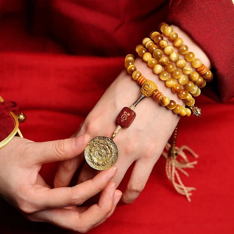 Layered Tiger Eye mala bracelets worn on two hands with carved wooden charm and brass medallion accent, Tibetan red robe backdrop