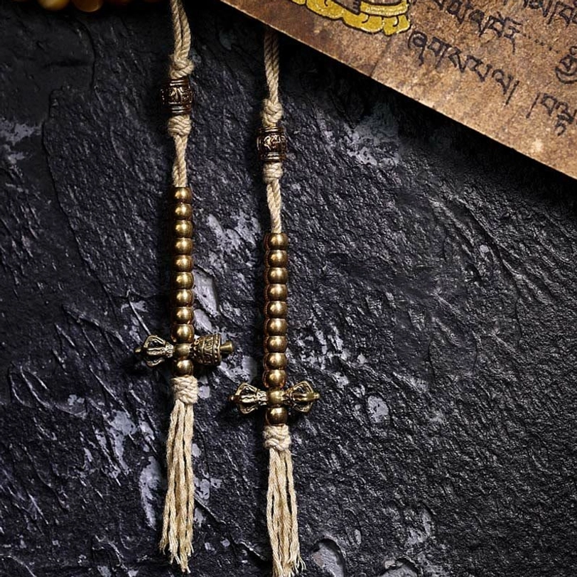 Mala tassel counters with copper Vajra charms, woven cotton tassels, and metal bead accents on dark stone and Tibetan script backdrop