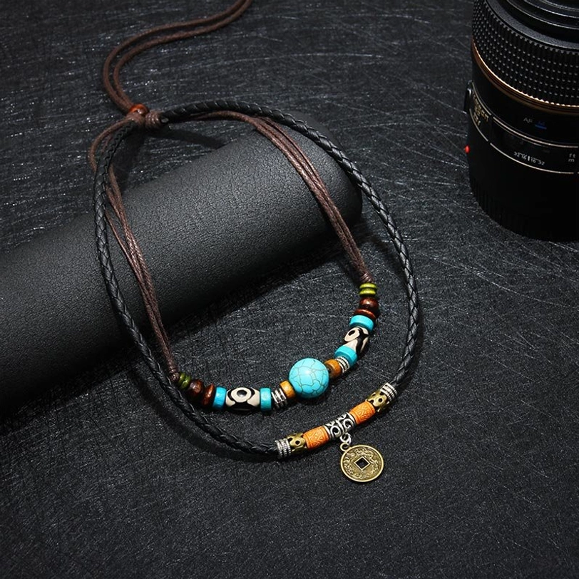 Close-up flat lay of turquoise Dzi bead protection necklace featuring dual leather strands and mixed tribal beads on dark fabric