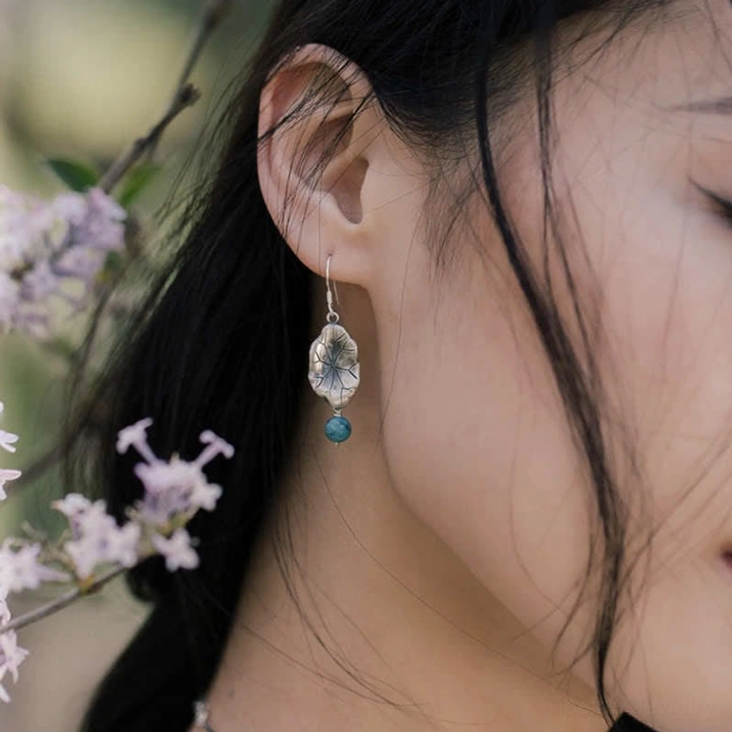 Ear close-up of female model wearing delicate turquoise lotus leaf earrings with lilac blooms background