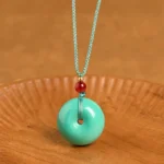 Turquoise peace buckle pendant necklace flat lay showing donut-shaped turquoise stone with teal woven cord and red accent bead on wooden background