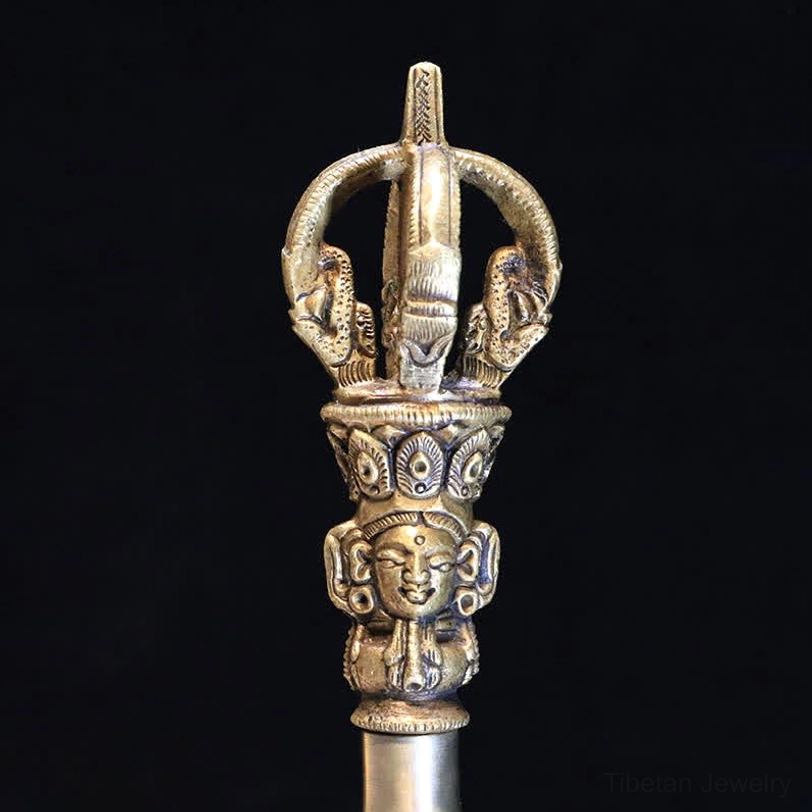 Vajra dorje five-pronged thunderbolt front close-up showing carved facial motifs and bronze patina finish on black background
