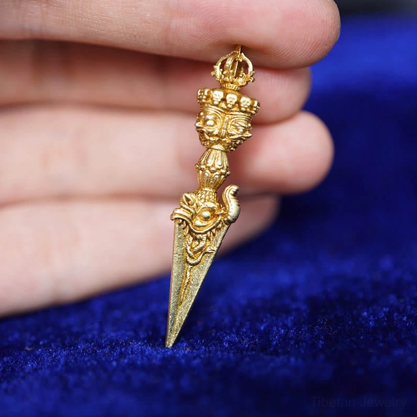 Tibetan Dorje Vajra held between fingers on blue velvet fabric, gold-toned finish with three-dimensional carved details and sacred geometry
