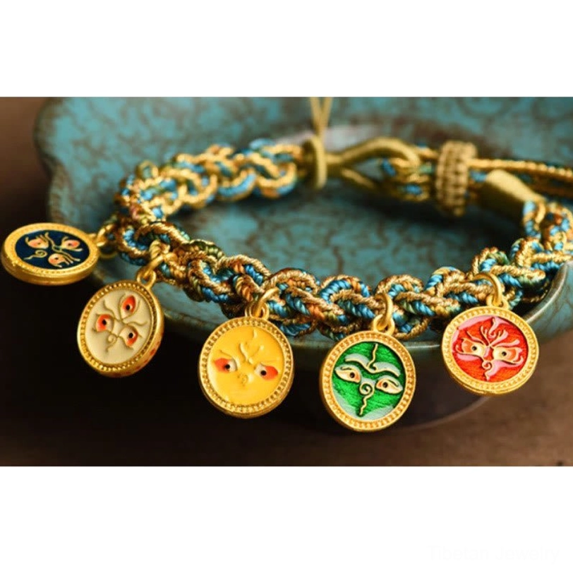 Detailed close-up of Tibetan charm bracelet with colorful enamel deity pendants and blue-gold twisted rope on ceramic tray background