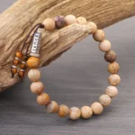 Weathered stone Om Mani Padme Hum bracelet with silver mantra charm on driftwood, overhead view showing earthy beads and tassel