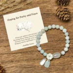 White jade purity bracelet with shell flower charm and blessing card on wooden table with natural decor - gift set flat lay