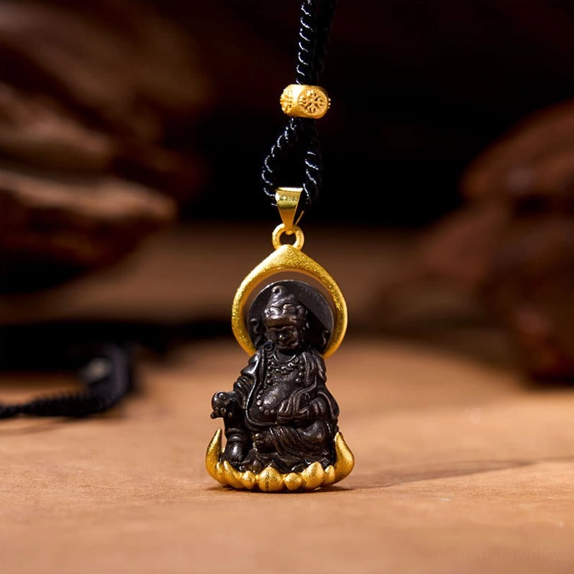 Yellow Jambhala Buddha pendant necklace frontal view, featuring dark metal Buddha statue seated on gold lotus base with protective halo, attached to black braided cord with gold decorative bead on wooden table