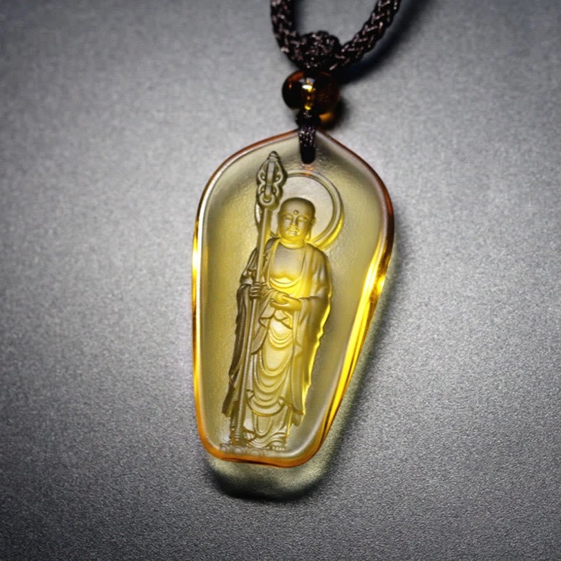 Amber yellow Ksitigarbha Buddha Liuli crystal pendant with elongated oval shape, standing Bodhisattva with halo and staff, black braided cord with amber bead accent