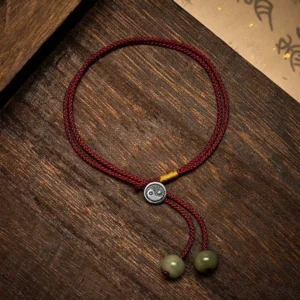 Overhead flat lay of Yin Yang protection bracelet showing braided red cord, 925 sterling silver charm, green stone beads, and golden cord accent detail on wooden table
