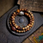 Tibetan Zakiram Goddess of Wealth bodhi seed mala bracelet with amber dzi beads and agate, displayed on carved dark tray with ancient Buddhist scriptures